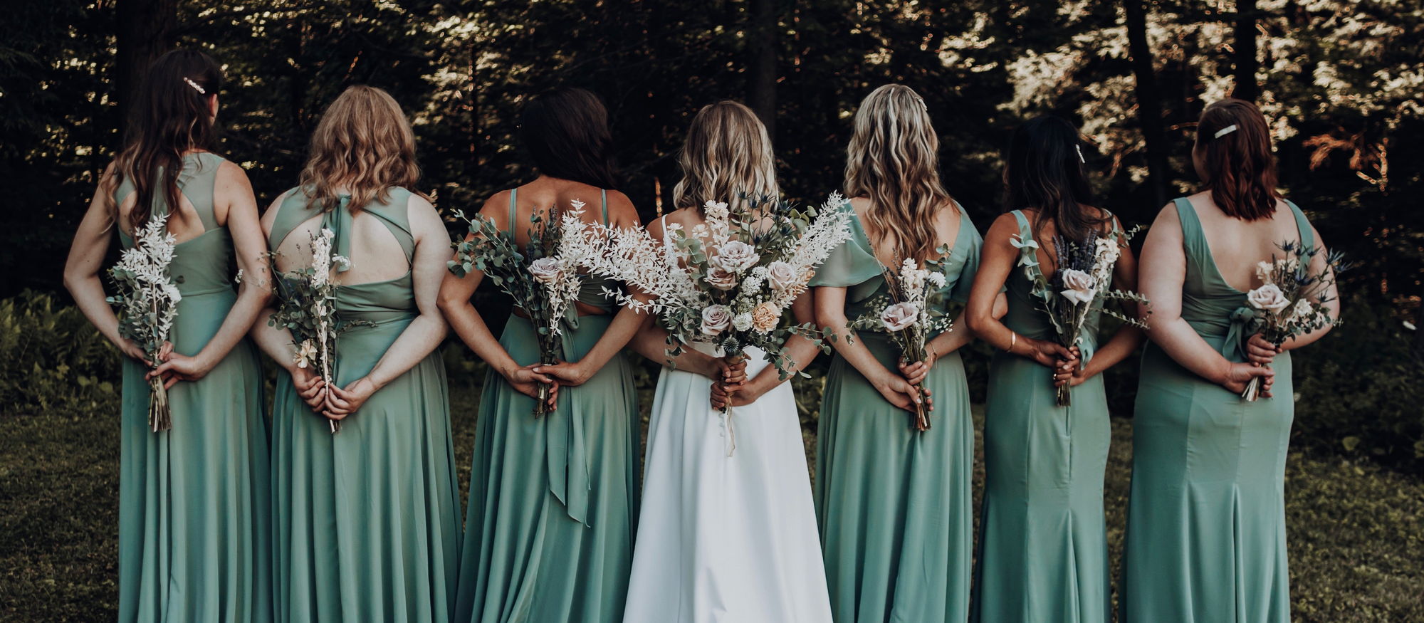 A wedding party in sage green seen from behind, each holding a bouquet of dried grasses, eucalyptus and blush roses